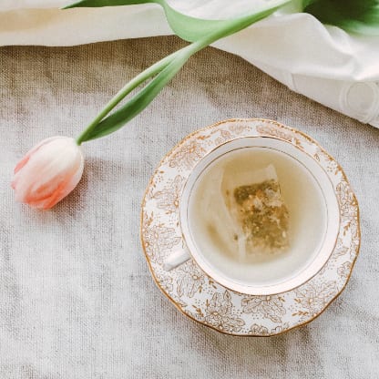 Teetasse mit goldverziertem Untersetzer und rosafarbener Tulpe auf Leinenstoff