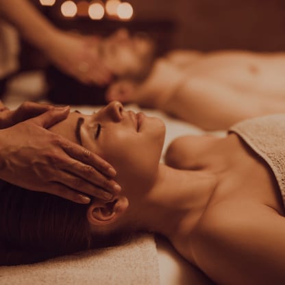 Sanfte Gesichtsmassage einer Frau bei Kerzenschein, ihr Partner entspannt im Hintergrund